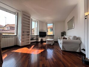 A modern living room with a wooden floor, a sofa, a television, and a window with a view of buildings. - CASA DEL POZZO (Rapallo)
