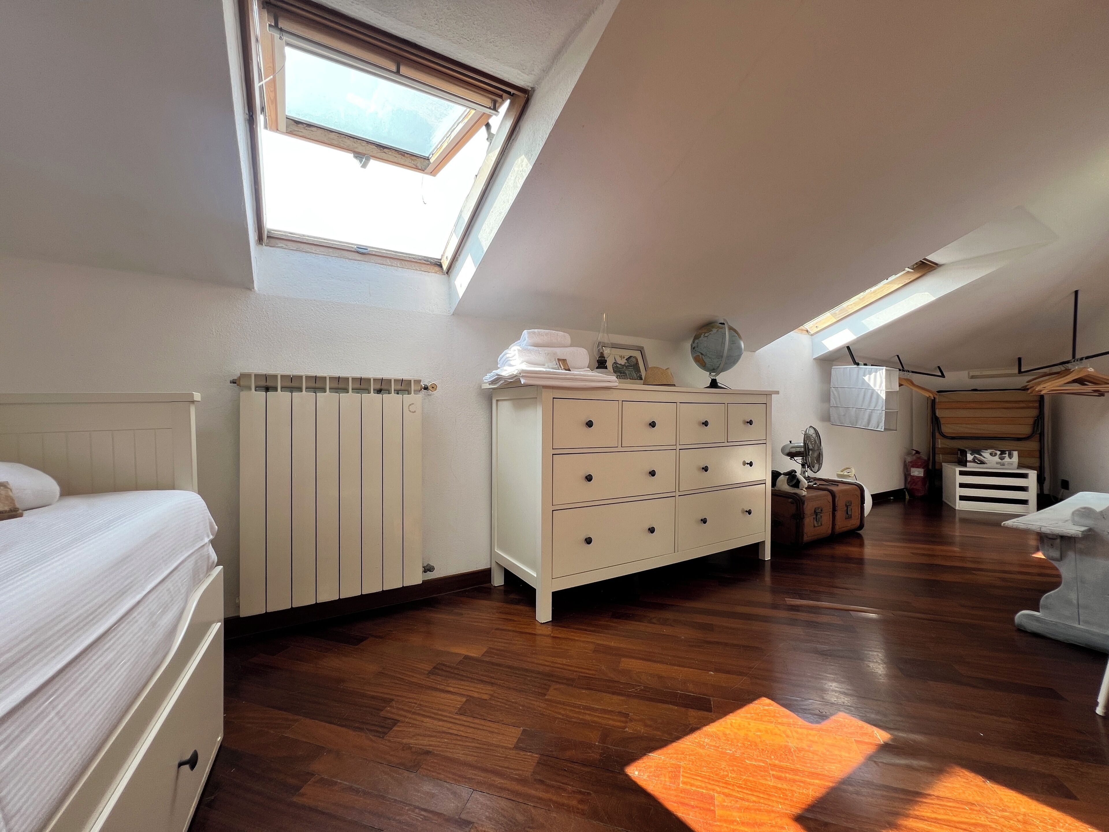 Un dormitorio con techo inclinado, radiador, cómoda, cama y tragaluz.