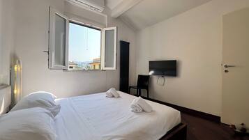 A hotel room with a bed, a flat-screen TV mounted on the wall, an open window, and an air conditioning unit.