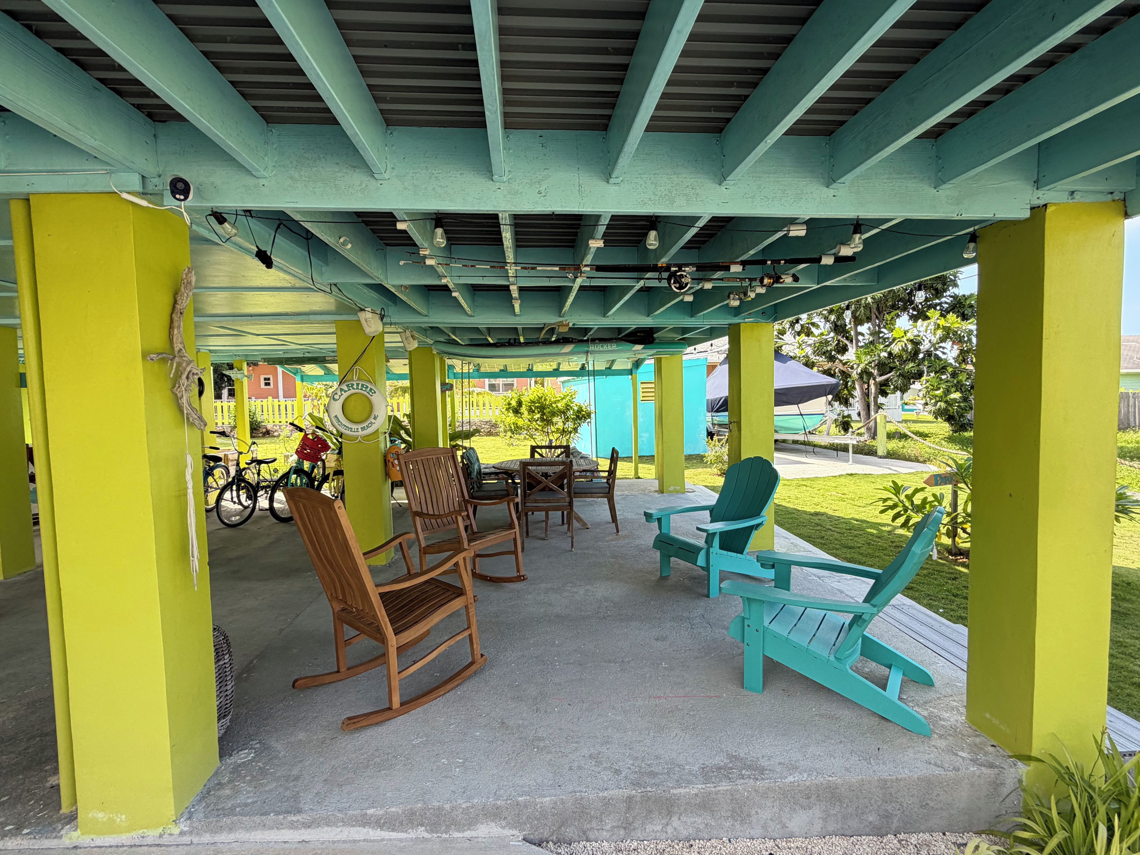 An outdoor patio with wooden rocking chairs and tables, a covered area with a green roof, and a view of a residential area in the background.