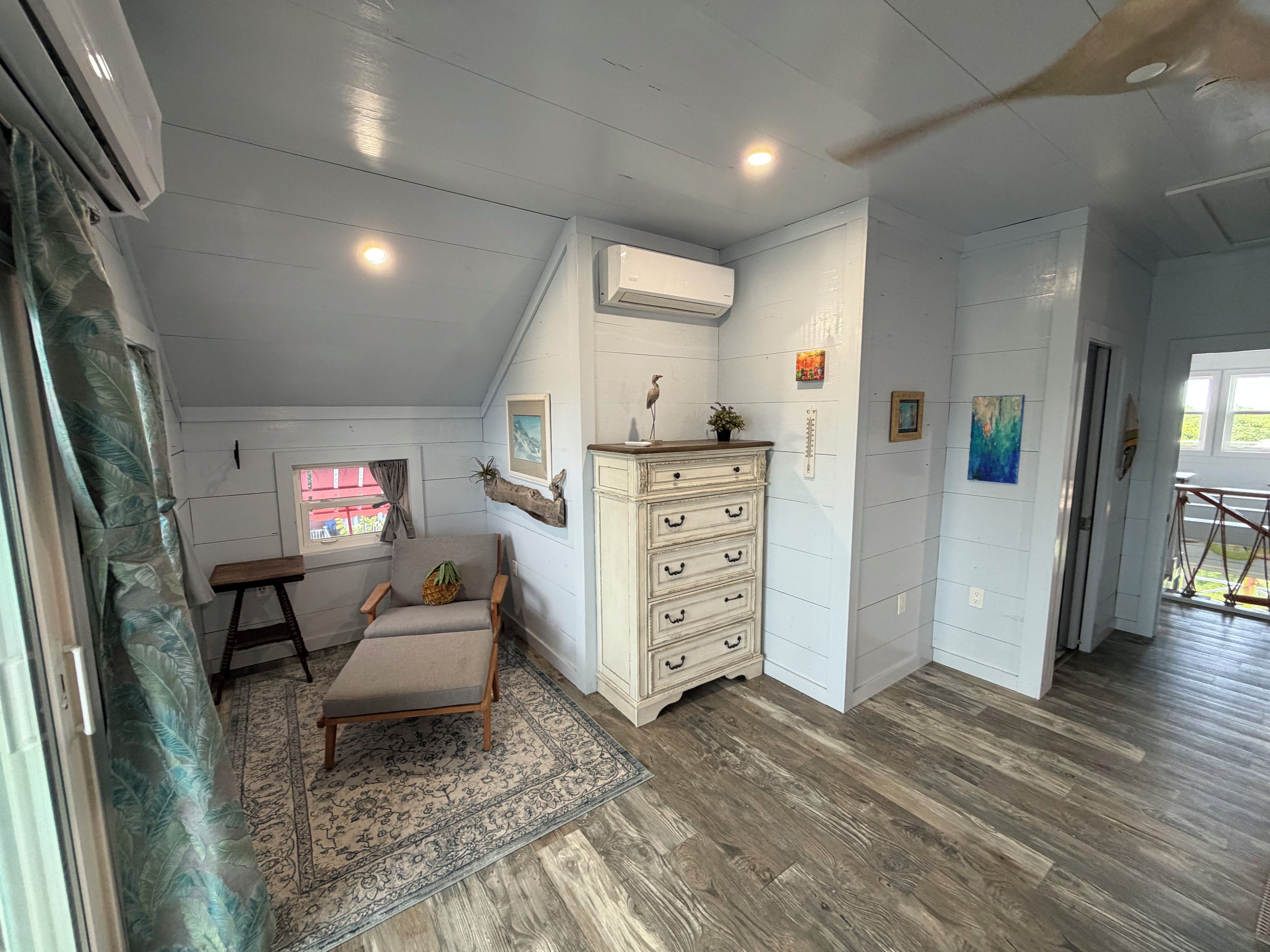A room with a wooden floor, a chair, a small table, a white dresser, and a window with a view.