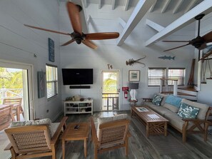 Un amplio salón con ventilador de techo, televisor de pantalla plana, mesa de centro de madera, mobiliario de mimbre y vistas al exterior.