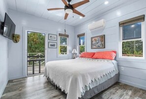 A bedroom with a bed, ceiling fan, air conditioning unit, and windows.