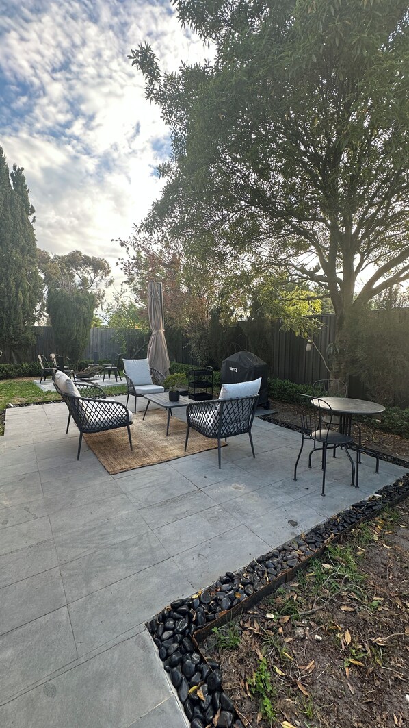 Un patio al aire libre con suelo de hormigón, rodeado por un bajo borde de piedra. Hay varias sillas y una mesa, con un árbol grande que proporciona sombra.