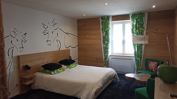 A bedroom with a wooden headboard, a bed with green and black pillows, a small round table, a green chair, and a wall with abstract cow drawings. - Beautiful Apartment in the Heart of Les Rousses Village (Les Rousses)
