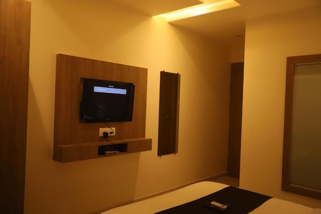 A hotel room with a mounted television, a small shelf below it, and a bed with a remote control on the bed.