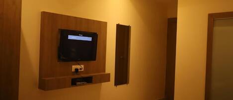 A hotel room with a mounted television, a small shelf below it, and a bed with a remote control on the bed.