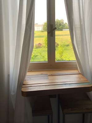Ikkunan edessä oleva puinen penkki, jonka takana on valkoiset verhot, ja josta avautuu näkymä vehreään maisemaan.