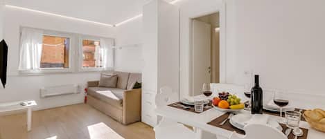 A modern, well-lit living area with a dining table set for two, a sofa, and a view of the kitchen through an open door.