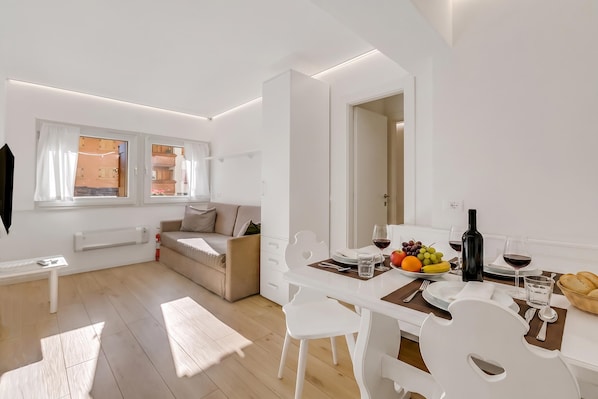 Sala de estar moderna e bem iluminada, com uma mesa de jantar posta para dois, um sofá e vista para a cozinha através de uma porta aberta.