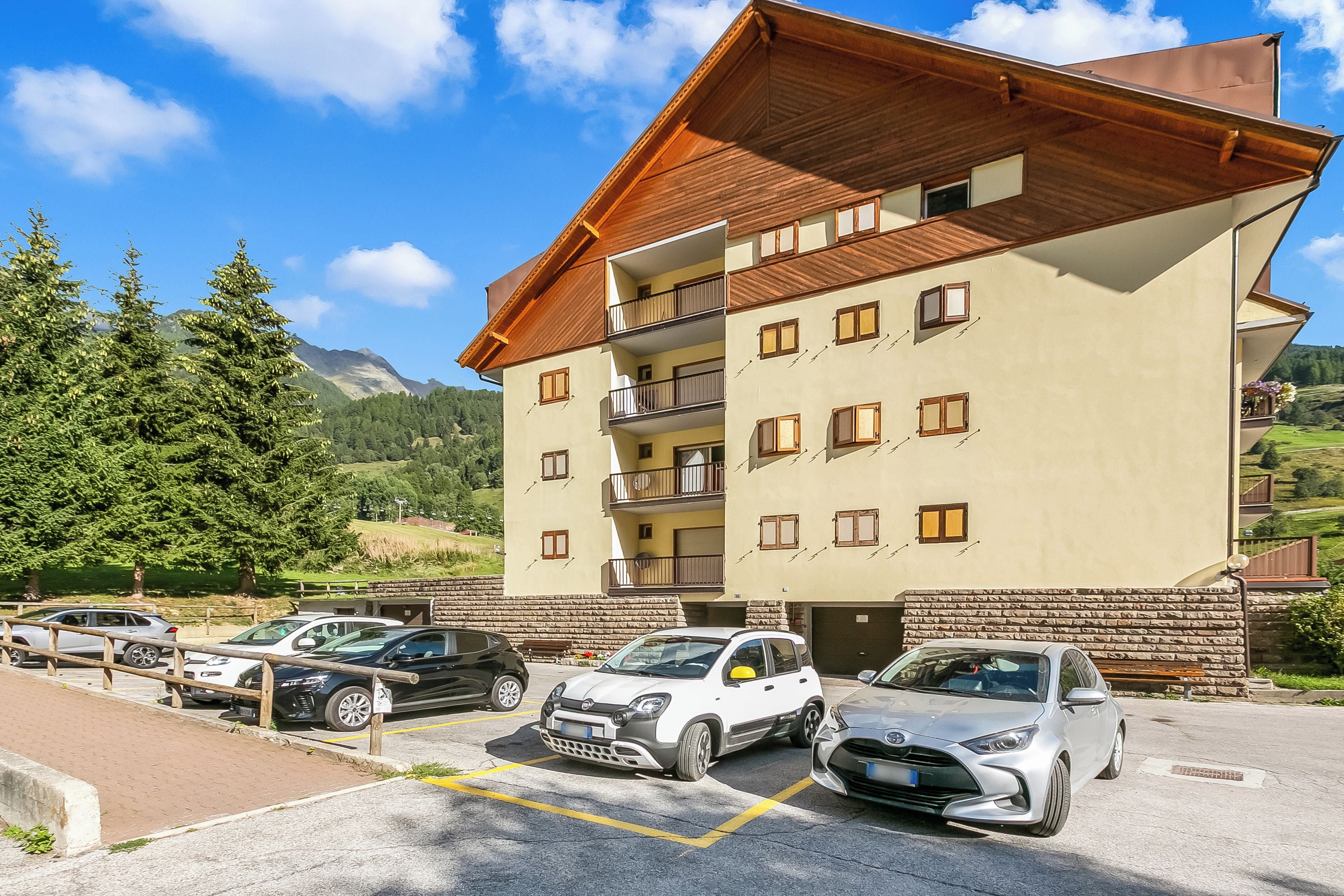 A multi-story hotel with a sloped roof, balconies, and a parking area with several cars.