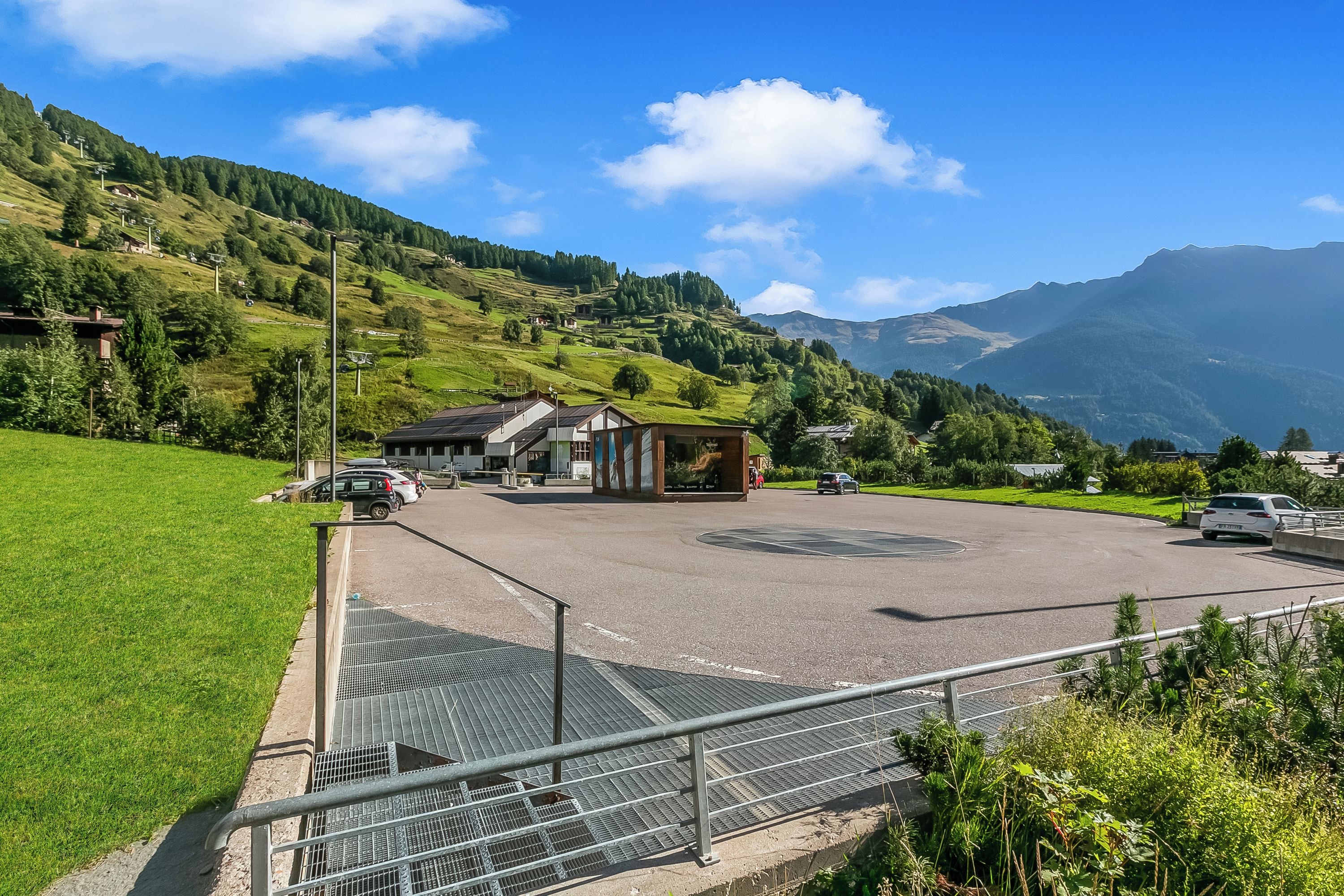 A parking area with a building in the background, surrounded by green hills and mountains.