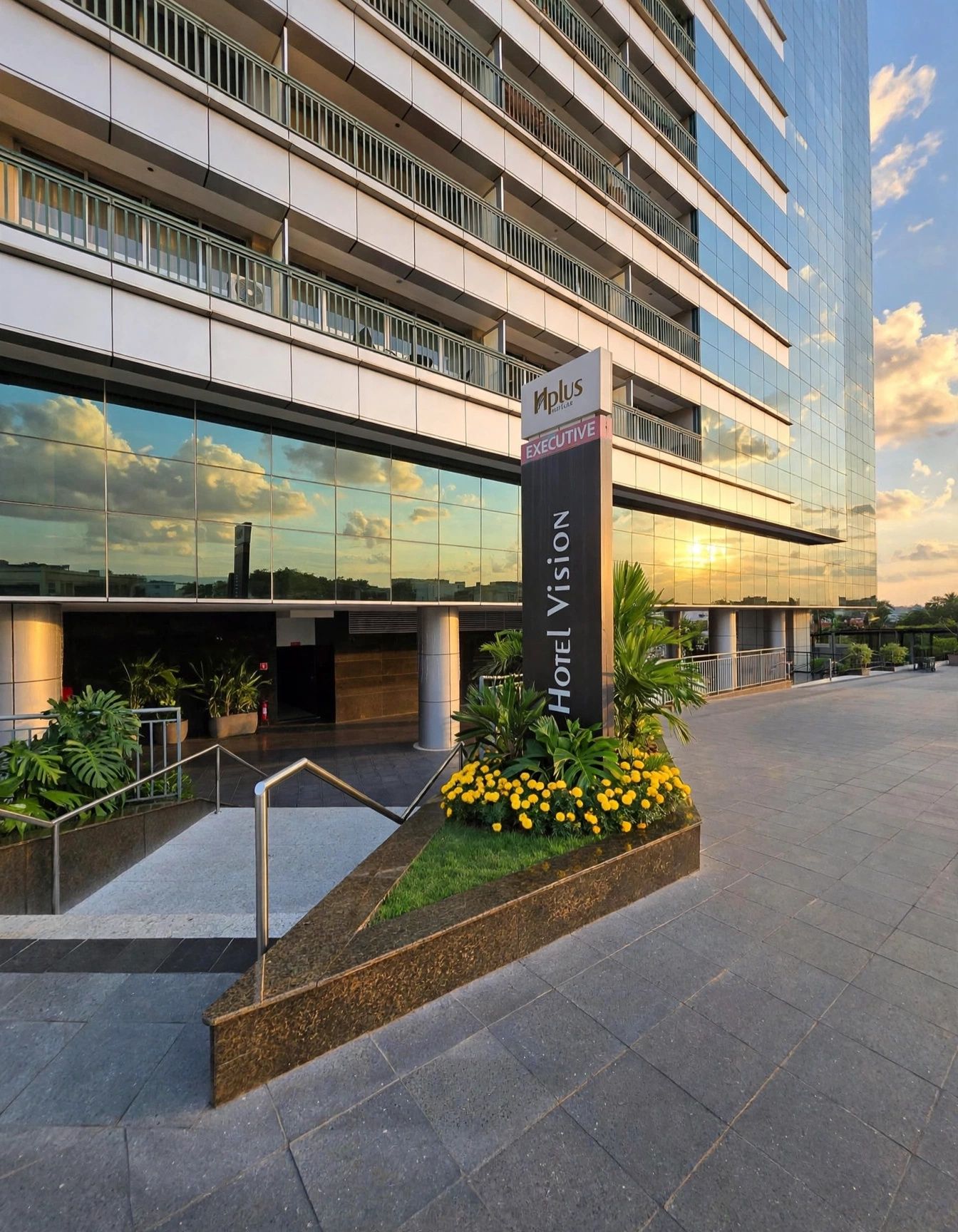 Um edifício moderno de hotel com fachada de vidro, um letreiro chamativo para 'Iplus Executive Hotel Vision' e uma área de entrada arborizada com plantas e um caminho.