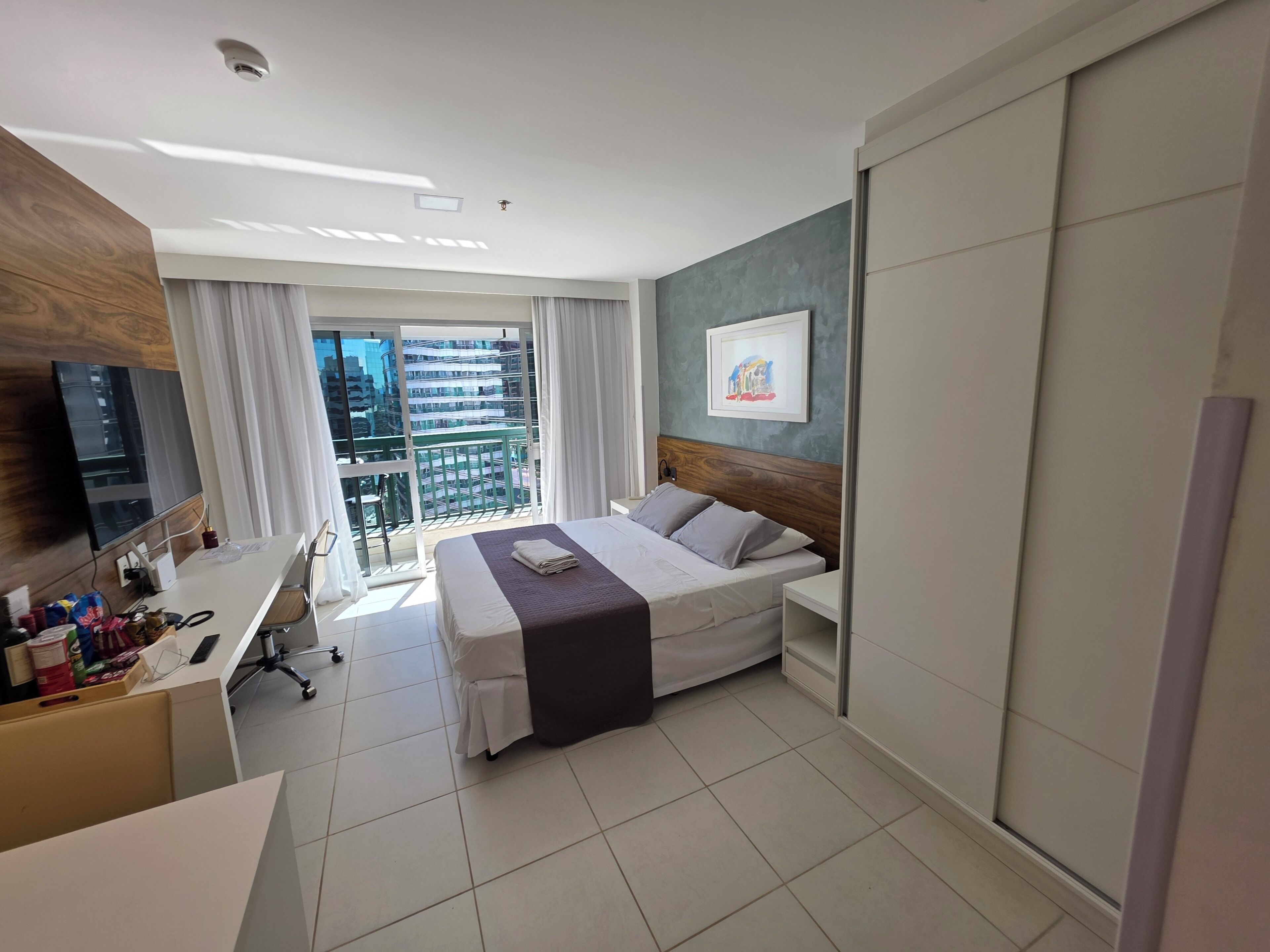 Habitación de hotel con cama, escritorio, silla, televisor, ventana con cortinas y balcón con vistas a edificios.