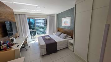Habitación de hotel con cama, escritorio, silla, televisor, ventana con cortinas y balcón con vistas a edificios.