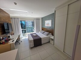 A hotel room with a bed, a desk, a chair, a TV, a window with curtains, and a balcony with a view of buildings.