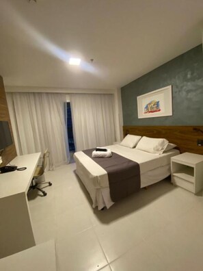 A hotel room with a bed, a desk, a chair, a bedside table, and a window with curtains.