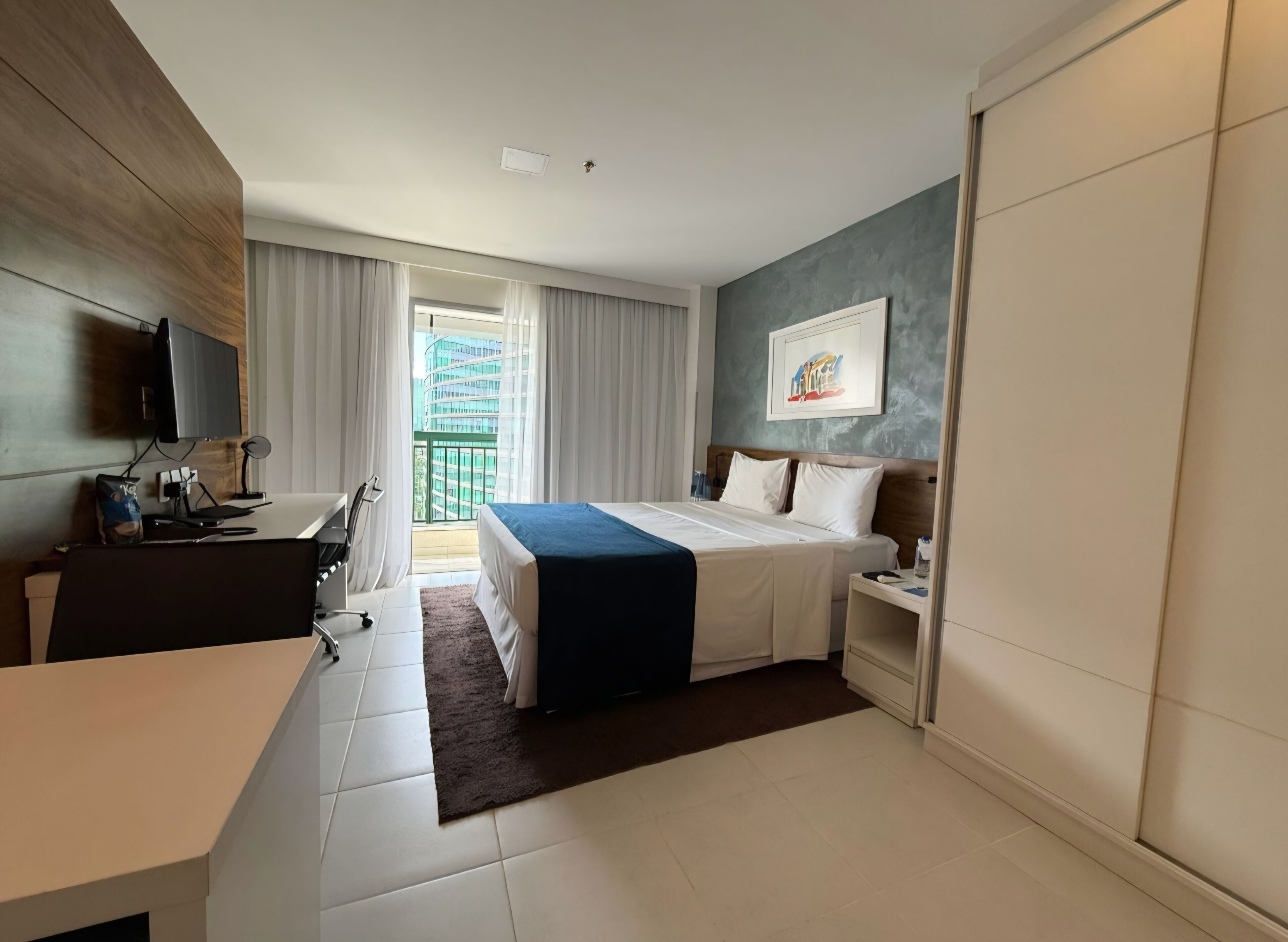 Una habitación de hotel con cama, escritorio, silla y vista a un edificio desde la ventana.