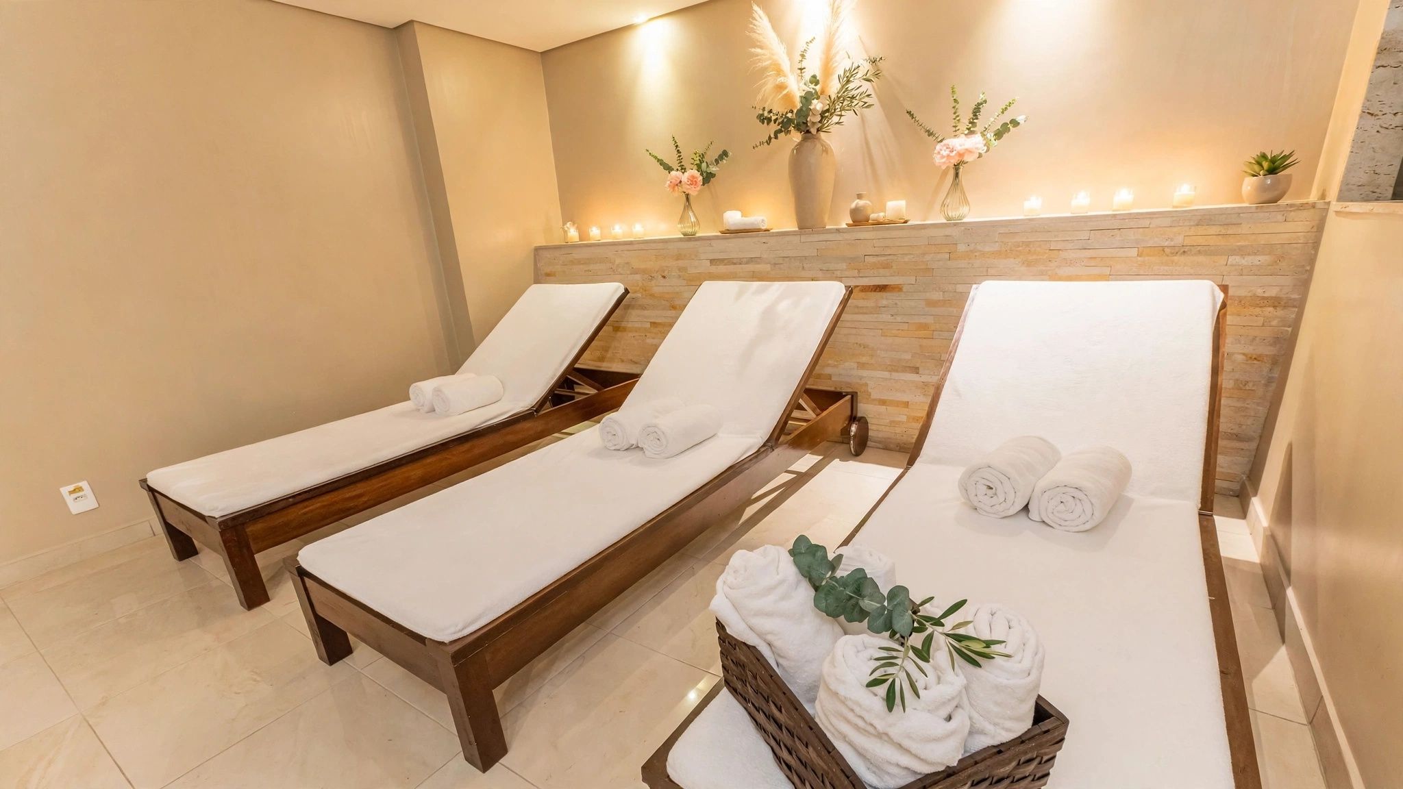 Un cuarto de spa con tres camastros de madera, un estante con velas y flores, y una cesta de mimbre con toallas.