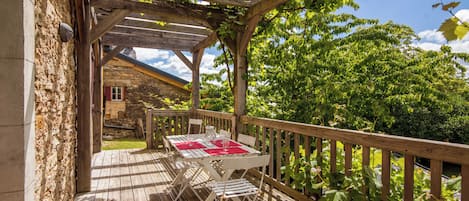 A wooden deck with a table and chairs, surrounded by greenery and a stone building.
