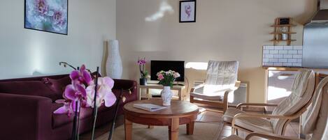 A living room with a sofa, a round table, a chair, a TV, and a decorative vase with flowers.