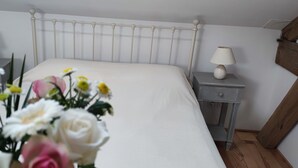 Ein Schlafzimmer mit einem weißen Bett, zwei Nachttischen mit Lampen, einem Holz-Kopfteil und einer Blumenkomposition auf der linken Seite.