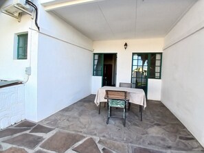 Un porche cubierto con mesa y sillas, suelo de piedra y una ventana con contraventanas verdes.