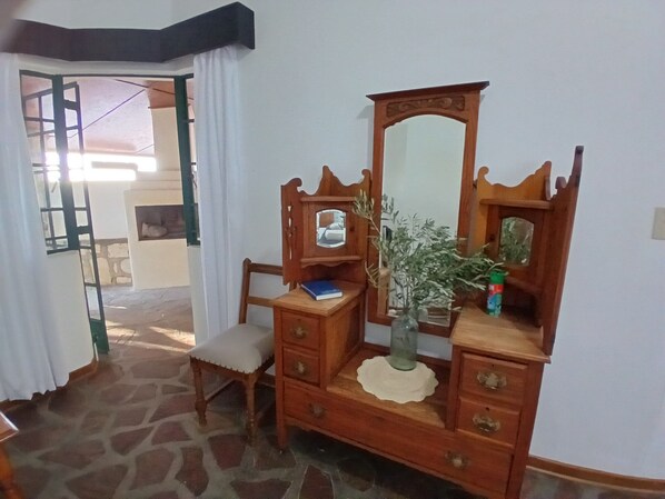 Una habitación con un aparador de madera, una silla, un espejo, un jarrón con una planta y un acceso a otra habitación.