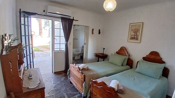 Quarto com duas camas de madeira, uma mesa com abajur, uma cadeira, luminária pendente e vista para área externa através de uma porta.