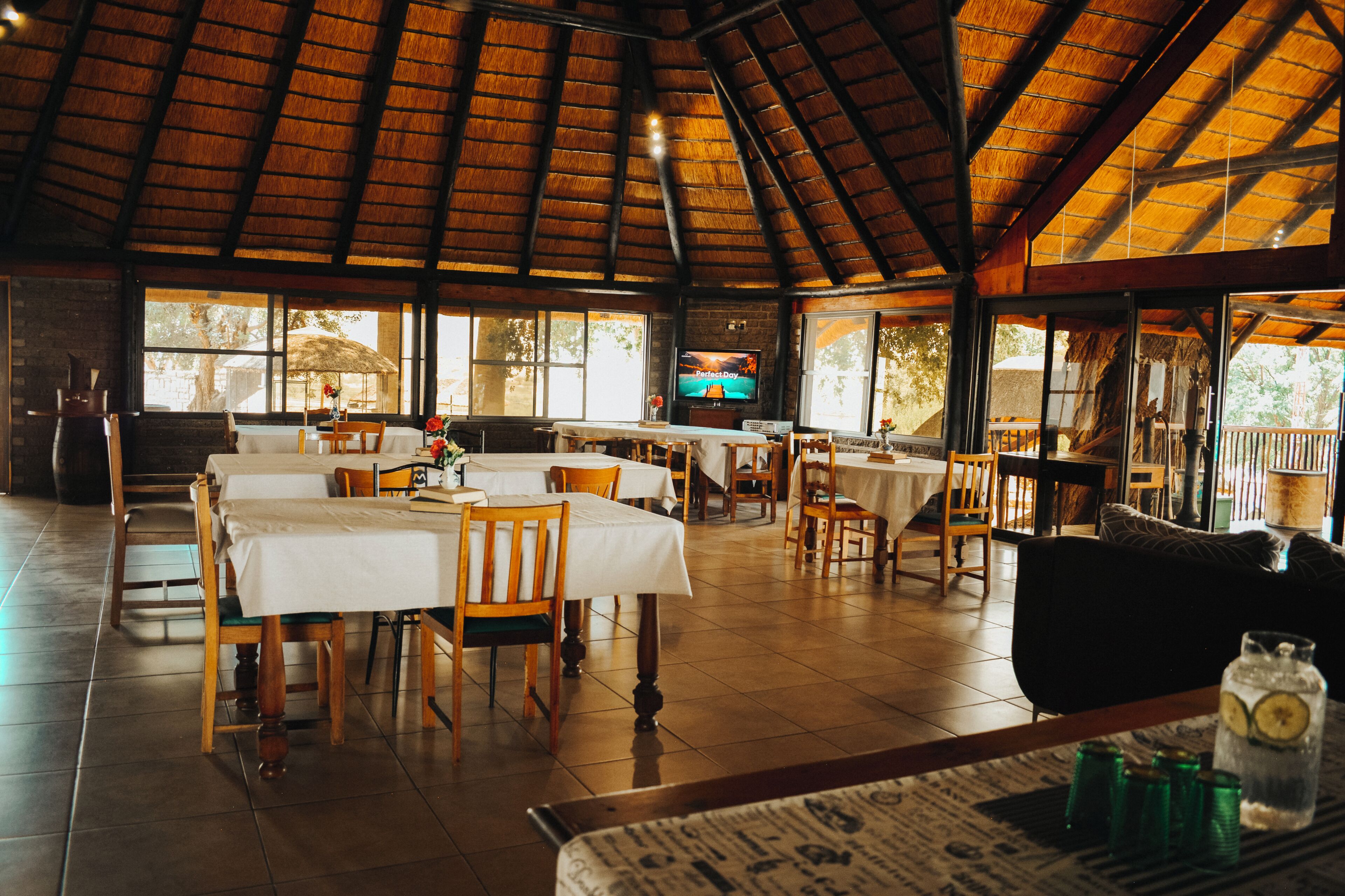 Un restaurante con varias mesas y sillas, una pantalla de televisión y un techo de paja.