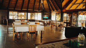 Um restaurante com várias mesas e cadeiras, uma tela de televisão e um telhado de palha.