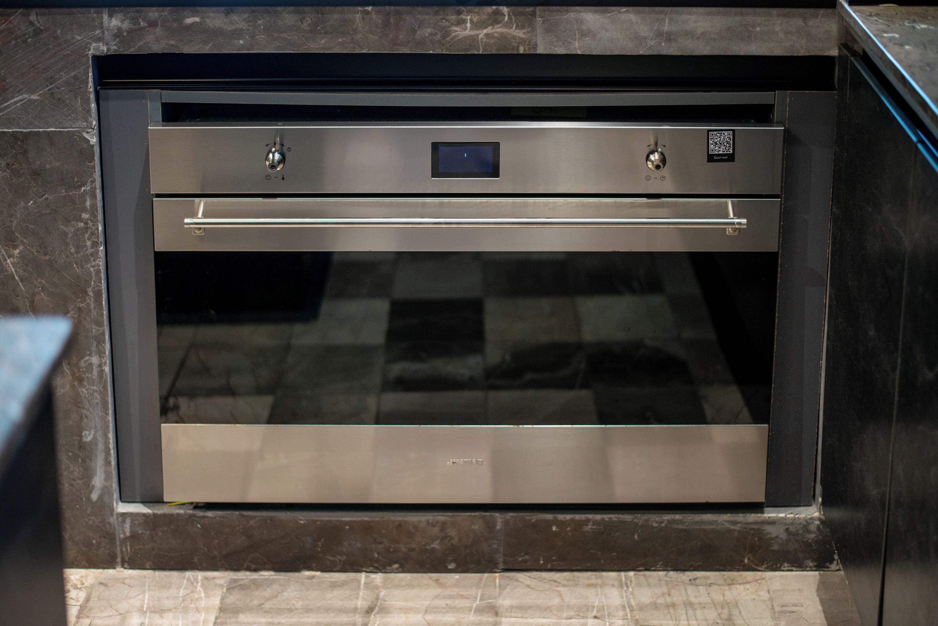 Forno incassato in acciaio inossidabile con display digitale e pannello di controllo, inserito in una cucina con piani di lavoro in marmo scuro.