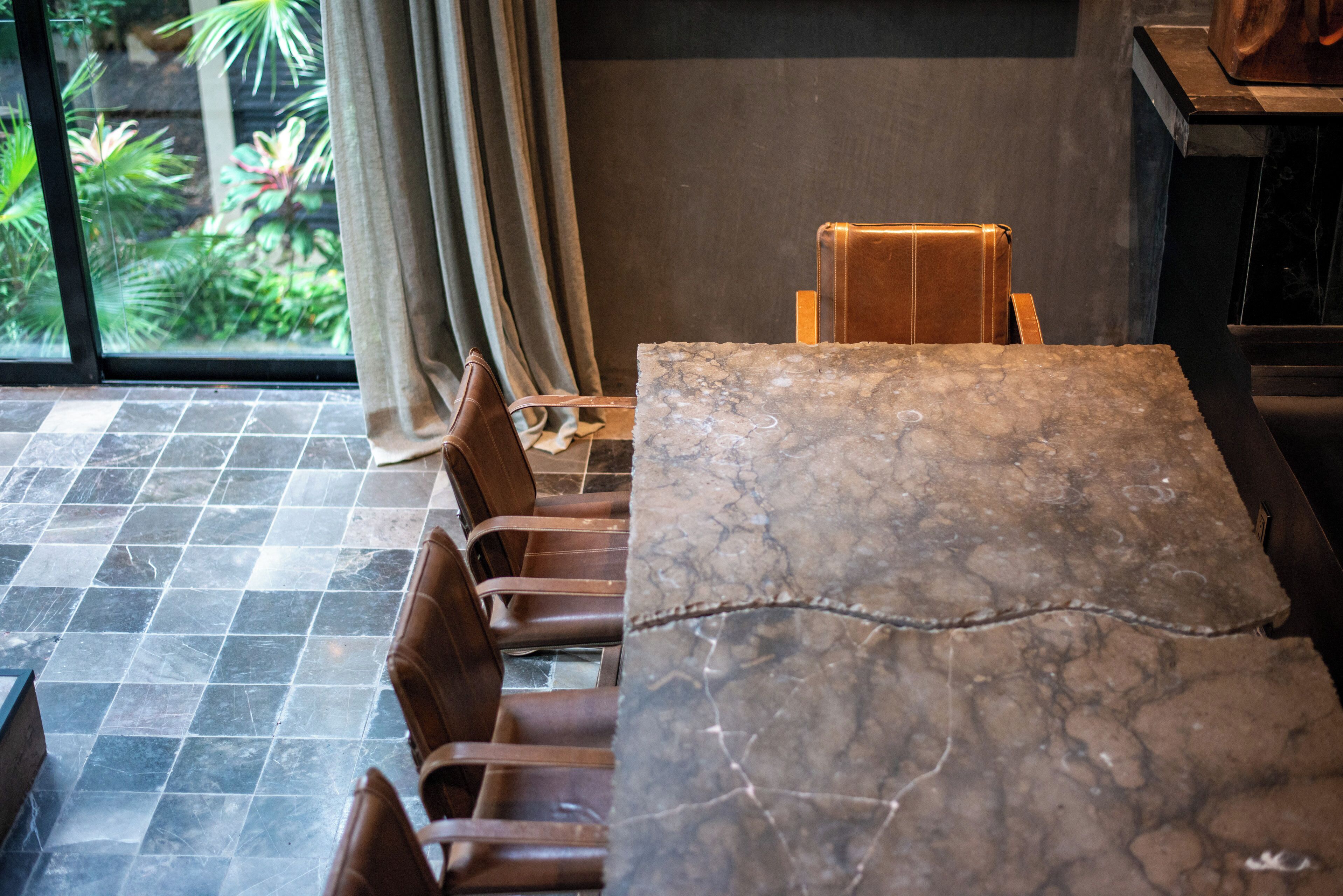 A dining area with a marble table and leather chairs, a view of greenery through a window, and a curtain.