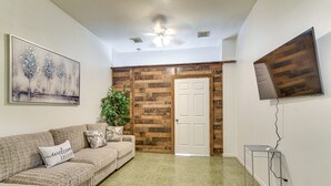 A living room with a sofa, a painting, a potted plant, a wooden door, and a TV mounted on the wall. - Inviting Unit, 10 Mi to Airport & Vintage Park! (Houston)