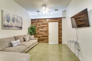 Ein Wohnzimmer mit einer Couch, einem Gemälde, einem Blumentopf, einer Holztür und einem an der Wand befestigten Fernseher.