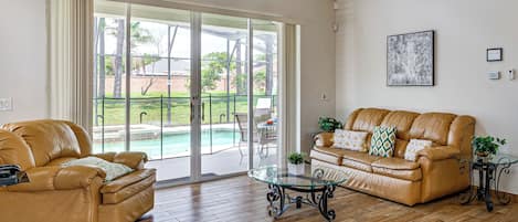 A spacious living room with a sliding glass door leading to a pool area, a leather sofa, a glass coffee table, and a framed artwork on the wall.
