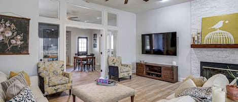 Amplia sala de estar con ventilador de techo, televisor de pantalla plana montado en la pared, chimenea y un comedor al fondo.