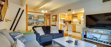 A living room with a sofa, a coffee table, and a television. There is a kitchen area with wooden cabinets and a refrigerator.