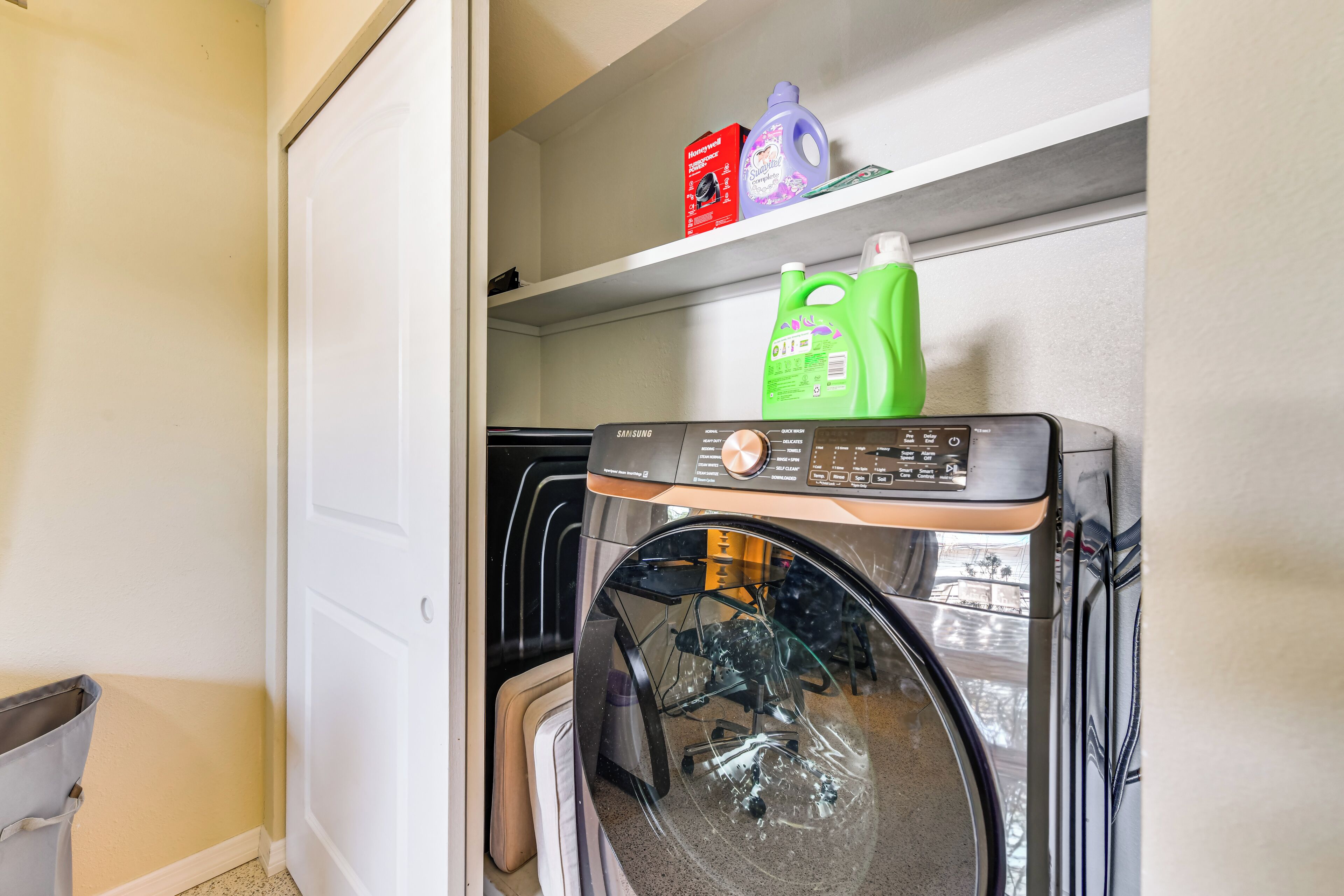 A laundry area with a front-loading washing machine, shelves with detergent bottles, and a door leading to another room.