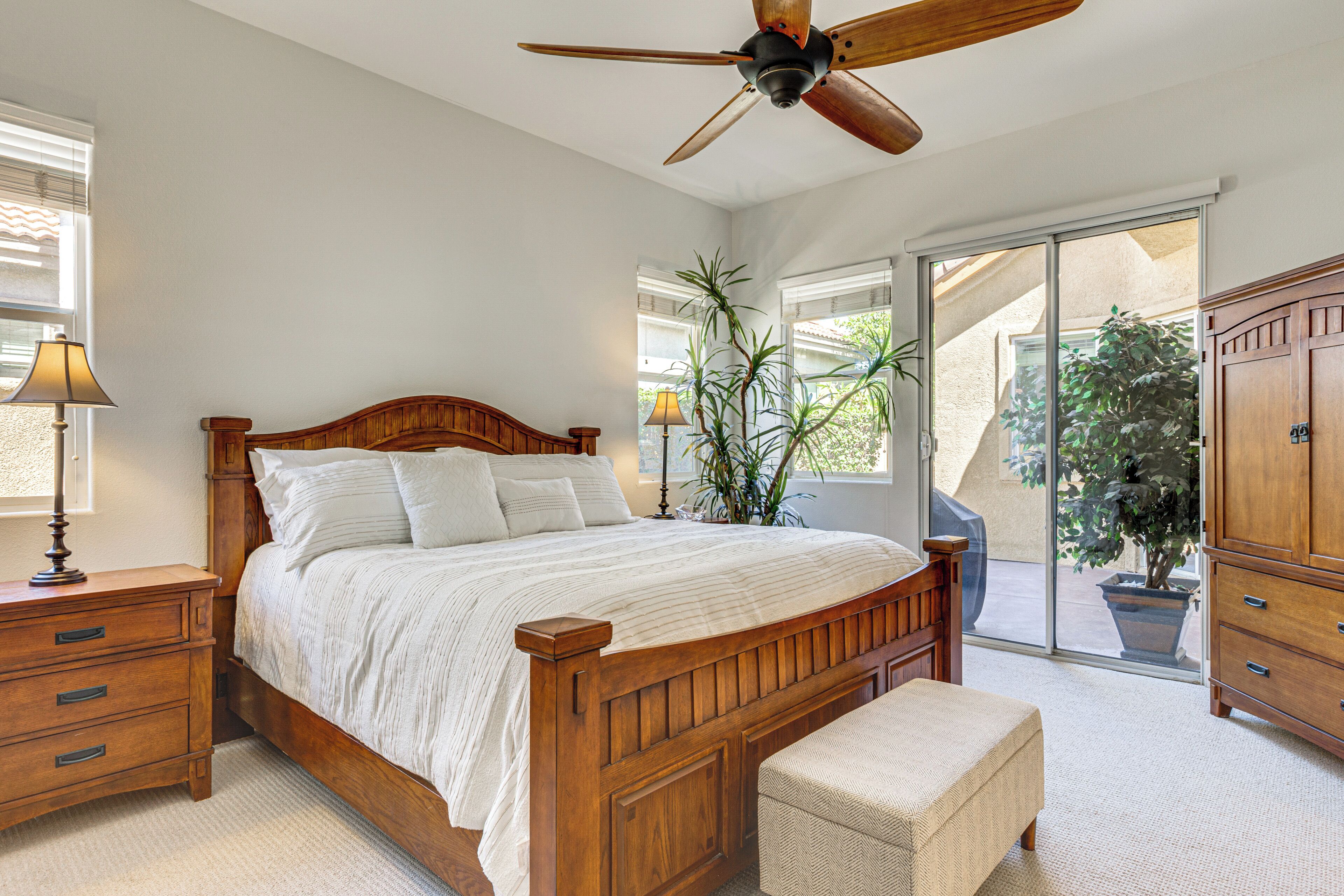 Une chambre avec un lit en bois, une table de chevet, une lampe, un ventilateur de plafond et une vue sur l’extérieur, accessible par une porte-fenêtre coulissante.