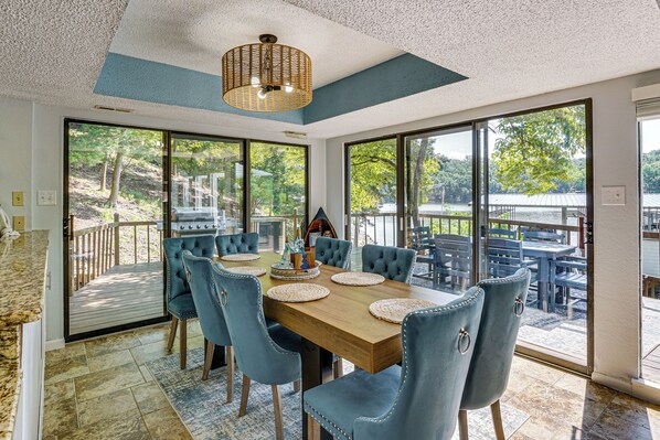A dining area with a wooden table and blue upholstered chairs, large windows with a view of trees and a body of water, and a ceiling with a textured finish.
