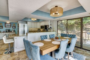 A modern kitchen with white cabinets, a wooden dining table with blue upholstered chairs, and a large window overlooking a forested area.