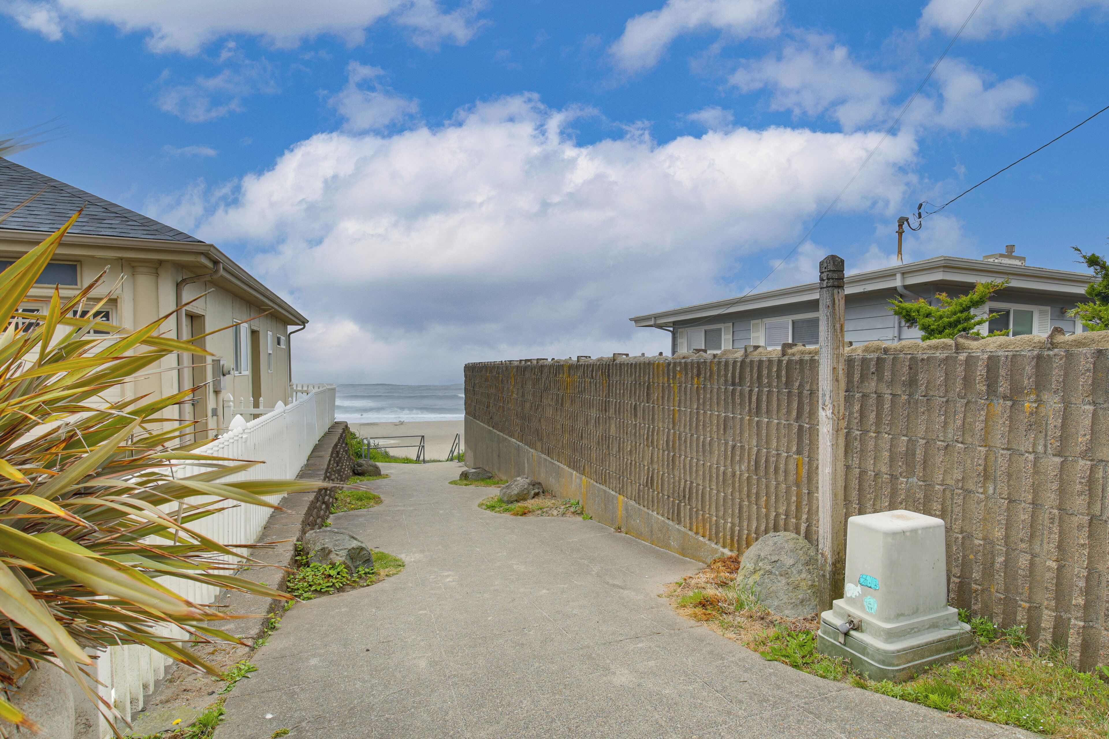 A coastal pathway leading to a beach, flanked by a wooden fence and a building with a white facade.