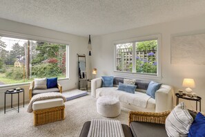 A cozy living room with a sofa, two armchairs, a coffee table, and a side table with a lamp. There are two windows offering a view of greenery outside. - Walk to Beaches! Lincoln City Family Home w/ Yard (Lincoln City)