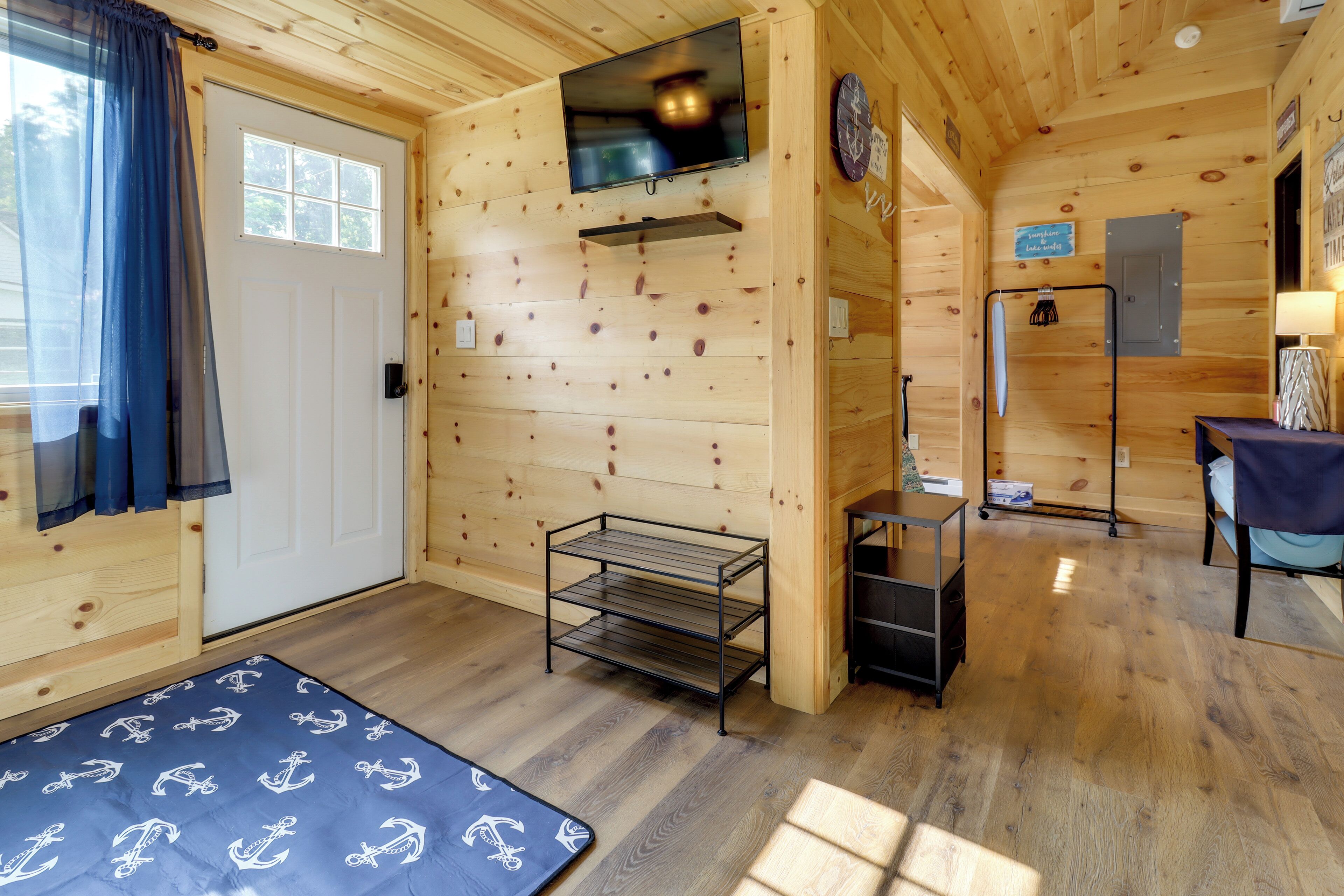 Interieur van een houten hutje met een aan de wand bevestigde televisie, een klein plankje, een rek en een vloermat met ankerprint.