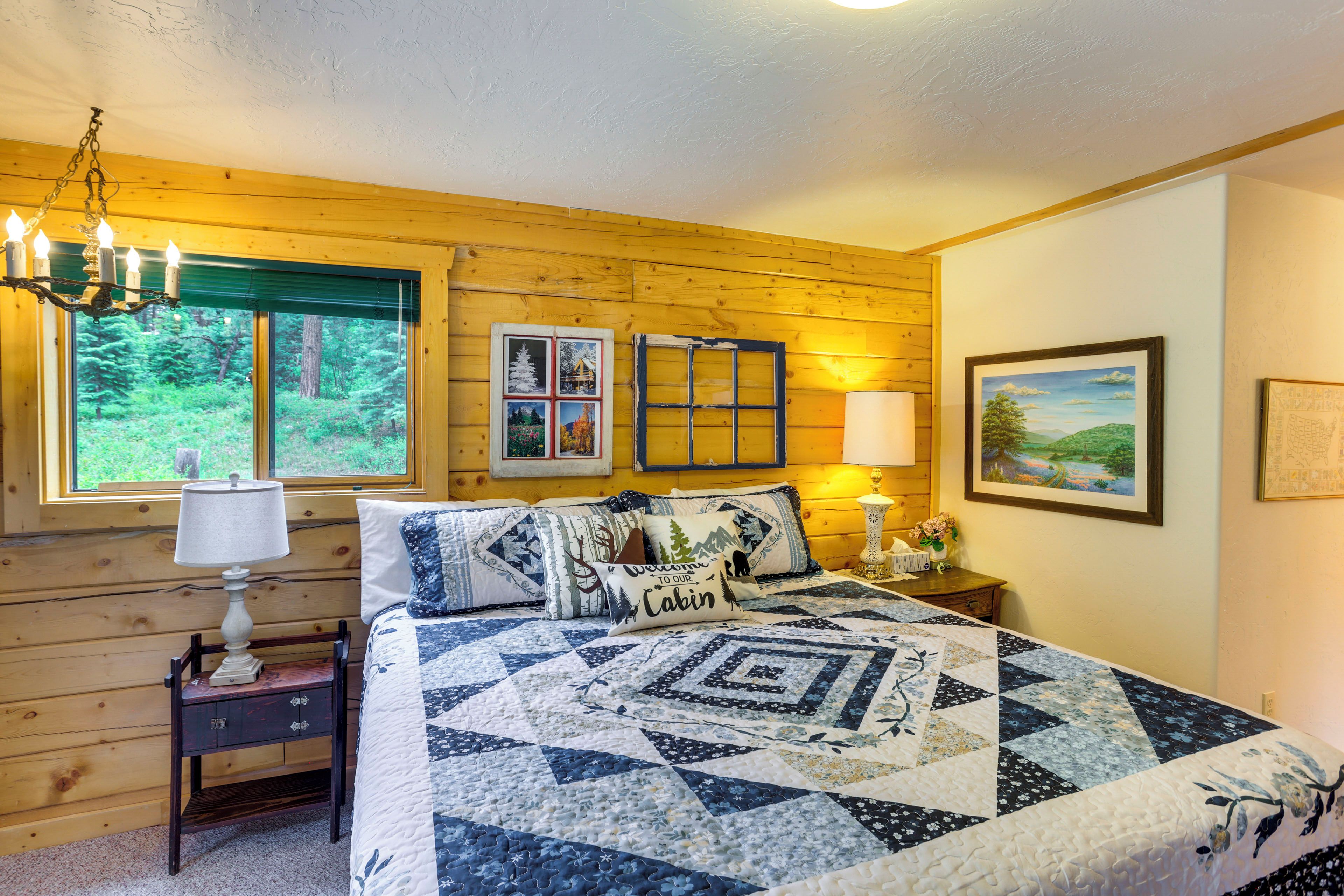 Um quarto aconchegante estilo cabana, com uma cama grande, uma mesa de cabeceira, um penteado, uma janela com vista para árvores e um quadro na parede.
