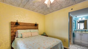 A bedroom with a wooden headboard, a bed with floral bedding, a nightstand, and a bathroom with a sink and mirror. - Pool, Pickleball & Beach! Gulf Island Club Condo (Hudson)