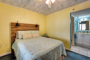 A bedroom with a wooden headboard, a bed with floral bedding, a nightstand, and a bathroom with a sink and mirror. - Pool, Pickleball & Beach! Gulf Island Club Condo (Hudson)