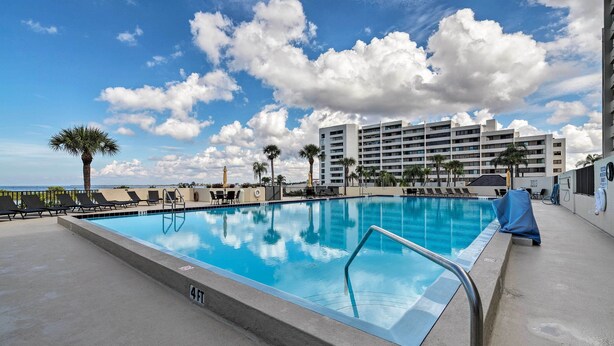 A swimming pool with a 4 FT depth marker, surrounded by lounge chairs and palm trees, with a hotel building in the background. - Pool, Pickleball & Beach! Gulf Island Club Condo (Hudson)