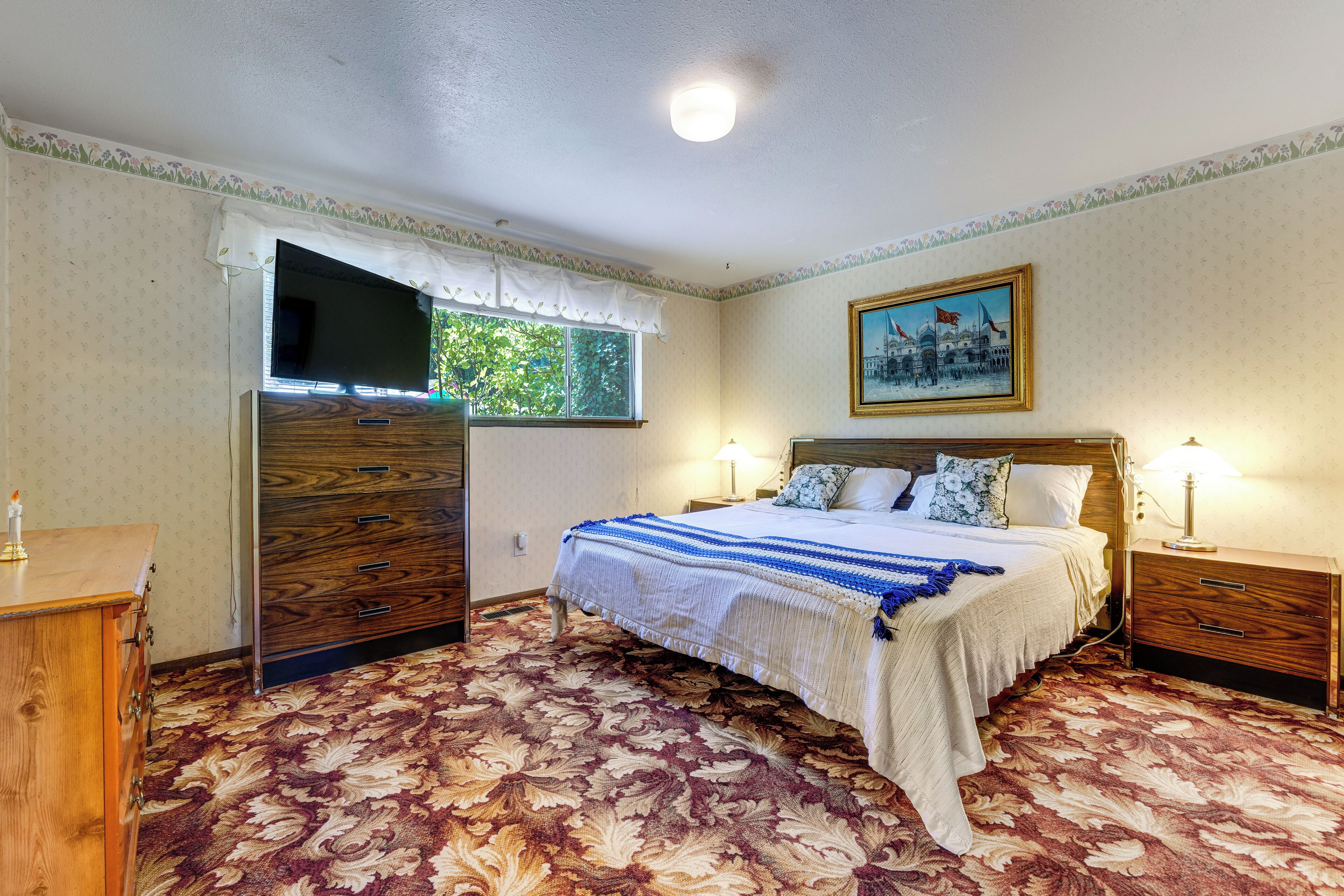 A bedroom with a bed, a nightstand, a dresser, a television, and a window.